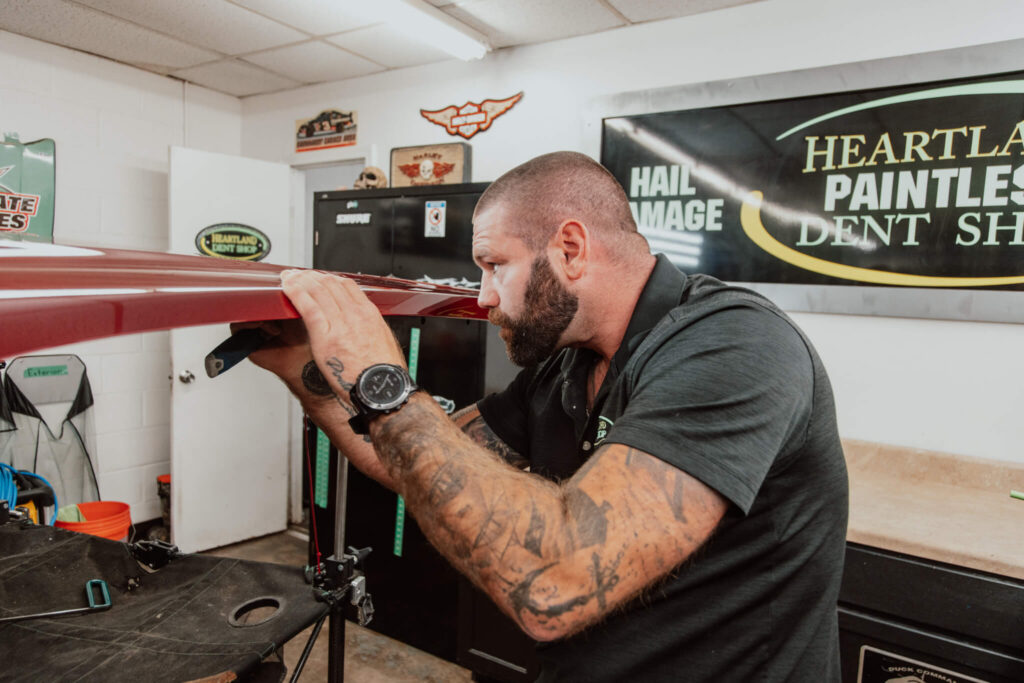 Heartland Dent Shop technician working a large dent out of a maroon panel with the shop banner behind him in Tulsa.