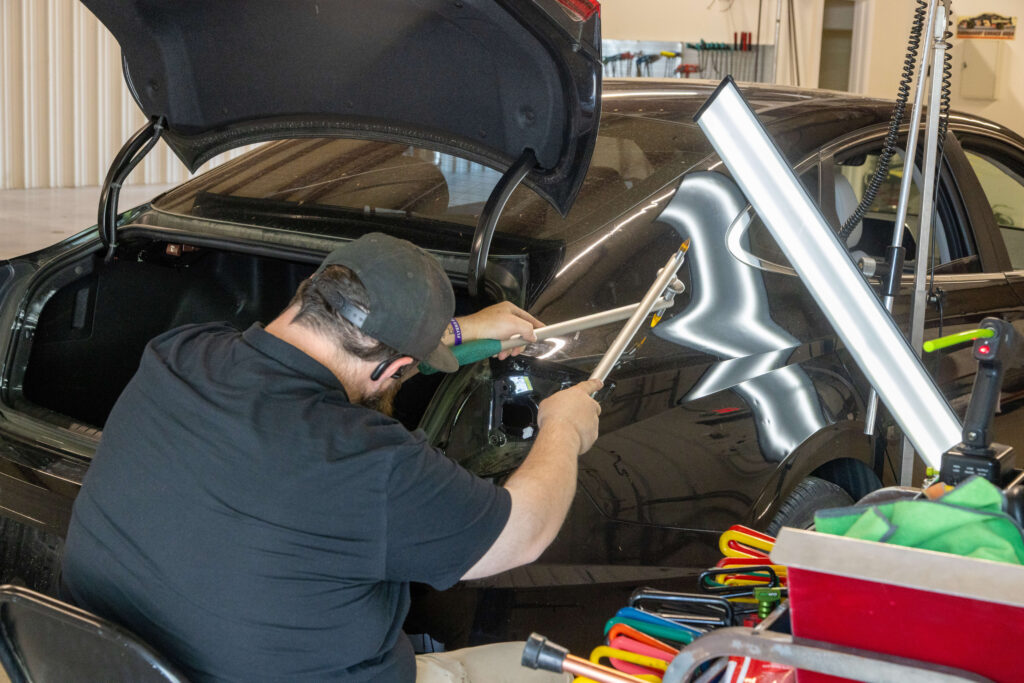 Heartland Dent Shop technician using a dent repair rod on a rear quarter panel with a lighted reflection board showing the damage.