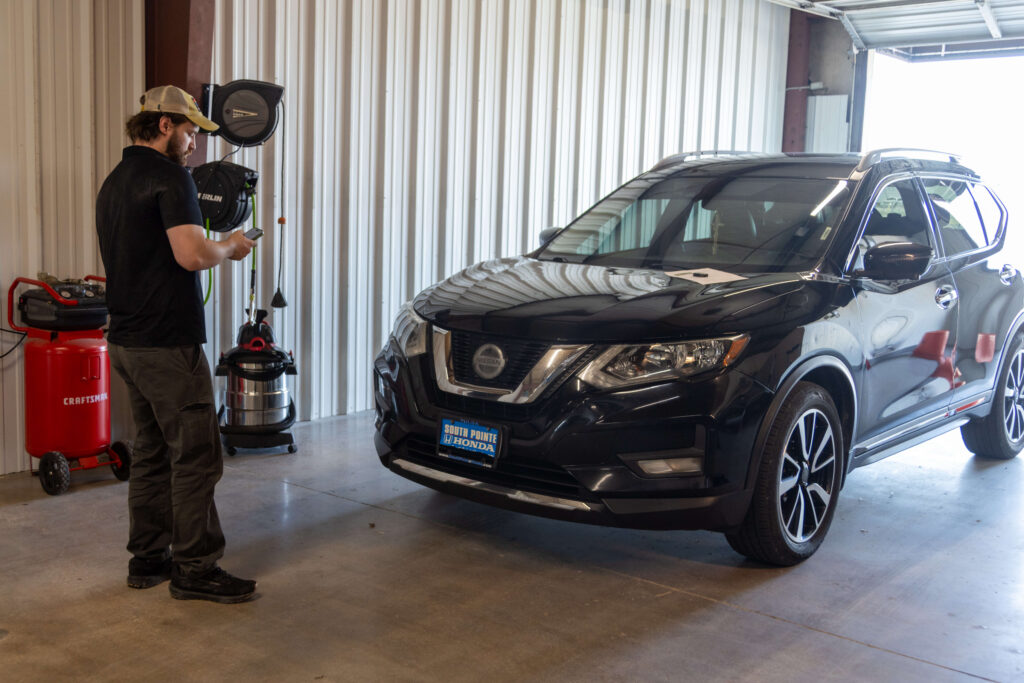 tech-documenting-damage-on-phone - Heartland Dent Shop Technician in a cap using a smartphone beside a black Nissan SUV in a metal-walled garage.